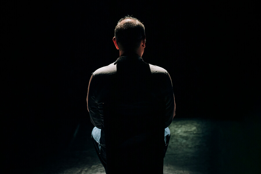 Silhouette of James Sutherland taken from behind. He is paritially lit, sitting on a chair on a theatre stage.