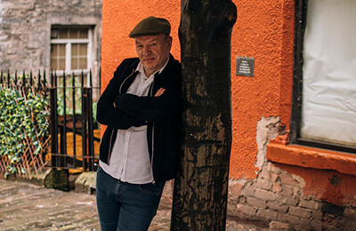 James Sutherland leaning against a tree in Old Edinburgh. He is looking at the camera, arms folded wearing a flat cap, white shirt and black jacket.