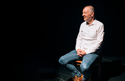 James Sutherland sat on a chair on a stage in a theatre, the background is black with just him lit by spotlight. James is miling, looking to the left.