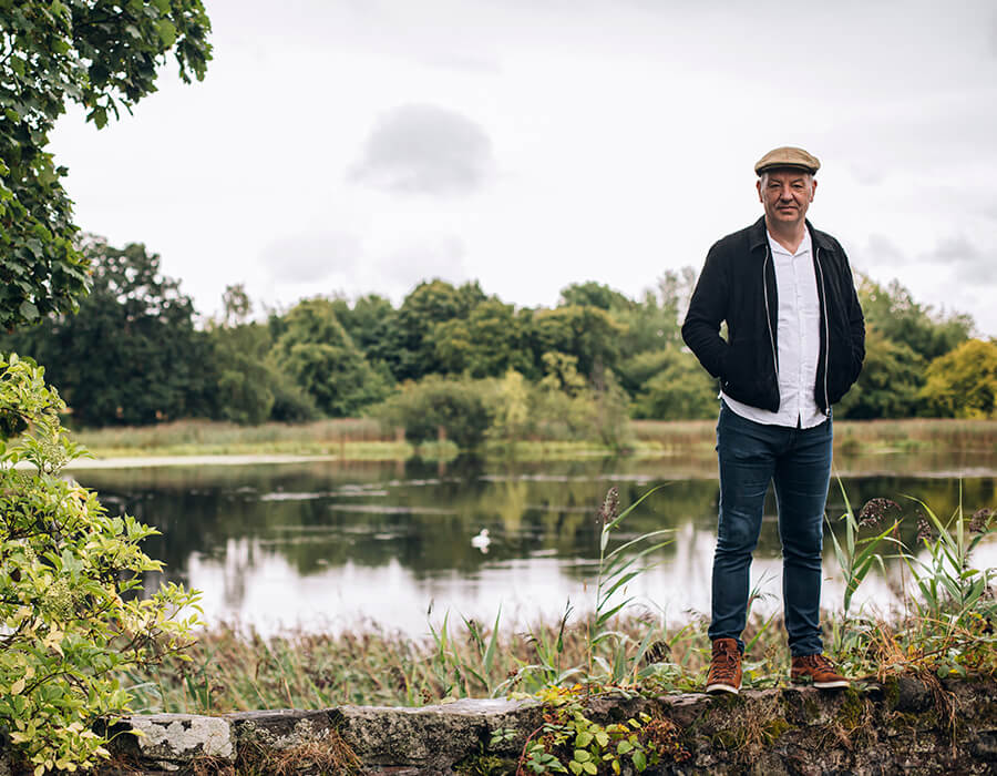 James Sutherland on an old brick wall with a lake, greenery and trees behind him.